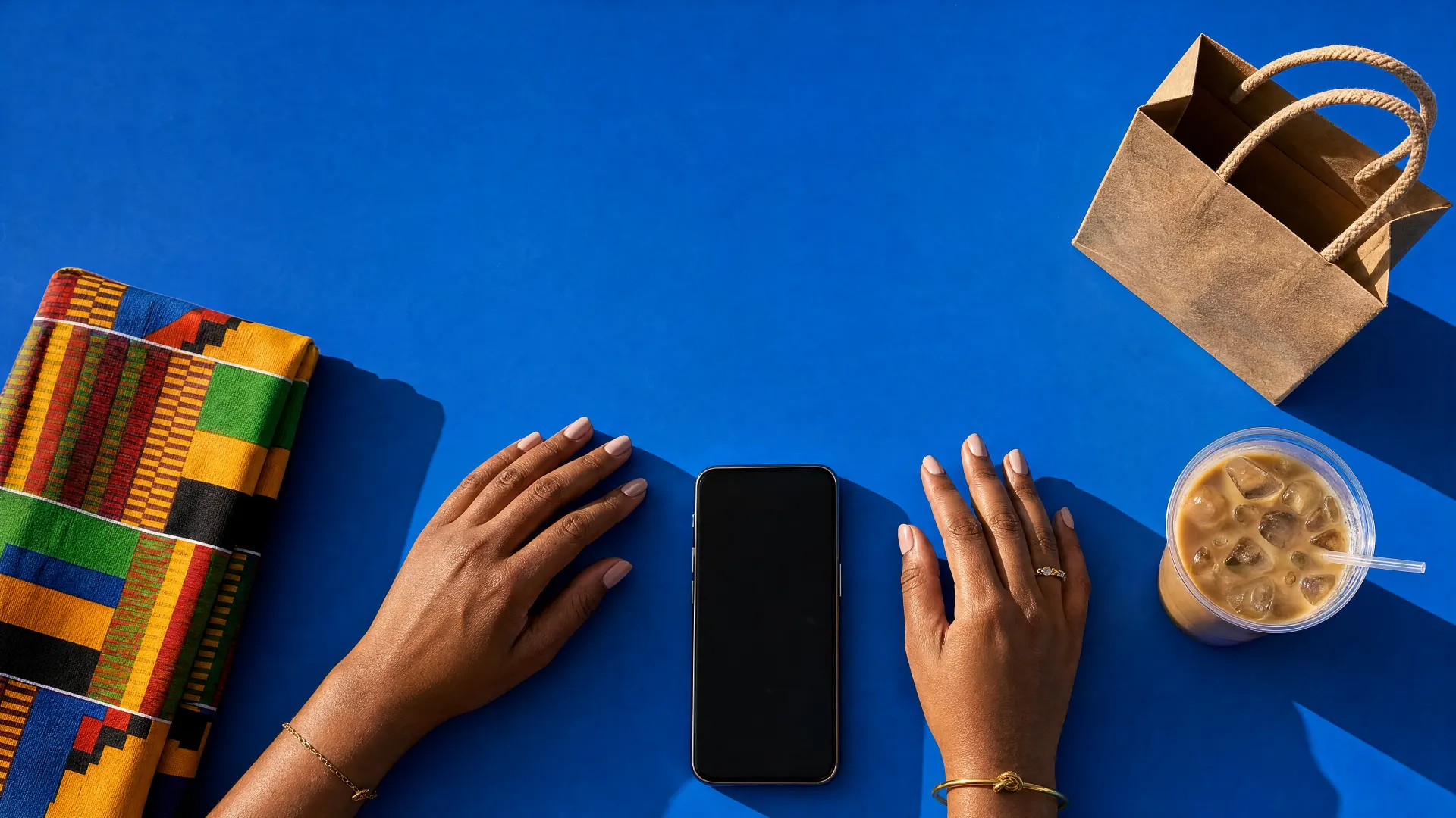 Hands resting either side of a phone on a blue table, beside kente cloth, a paper bag and an iced coffee